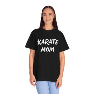 May include: Black t-shirt with the white text "KARATE MOM". The shirt is a short-sleeved, crew neck style. The text has a distressed, splattered effect. The person in the image is wearing light blue jeans.
