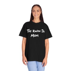 May include: Black t-shirt with the white text "Tae Kwon Do Mom". The shirt is a short-sleeved, crew neck style. The text is in a handwritten style font.
