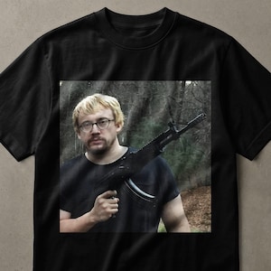 Peut inclure: Un t-shirt noir à col rond avec une impression graphique carrée sur la poitrine. L'image montre une personne aux cheveux blonds portant des lunettes et tenant un fusil noir dans une zone boisée. Le t-shirt a des manches courtes et une coupe standard.