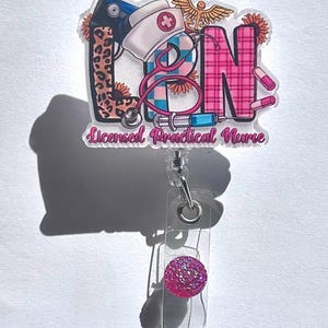 May include: A Licensed Practical Nurse badge reel with a clear plastic base and a pink glitter accent. The badge features the letters "LPN" with medical-themed illustrations, including a stethoscope, pills, and a nurse's cap. The background includes a leopard print and plaid pattern.