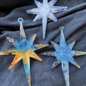 May include: Three eight-pointed star ornaments made of resin. One is white with blue glitter, one features a blue and orange gradient with gold glitter, and one is blue with multi-colored glitter. Each star has a small loop at the top for hanging.