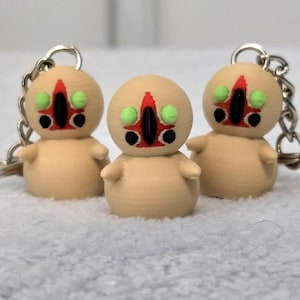 Op de afbeelding: Drie kleine, beige beeldjes met sleutelhangers. Elke figuur heeft een kenmerkend gezicht met rode, zwarte en groene details. De figuren staan op een wit, getextureerd oppervlak.