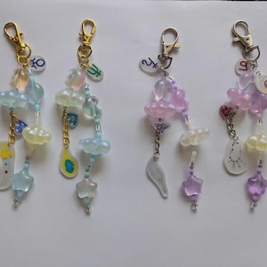 May include: Four handmade keychains with gold-colored clasps. Each keychain features a cascade of translucent beads in pastel colors, including blue, yellow, and purple. Decorative charms include stars, clouds, and wing shapes. The keychains are arranged on a white surface.