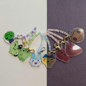 May include: A collection of handmade charms with cartoon character designs. Each charm is attached to a beaded chain with a gold-colored clasp. The charms feature various colors, including green, blue, yellow, and red, with white and black accents.