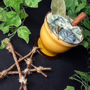 Peut inclure: Une bougie jaune dans un verre avec un bâton de cannelle, une feuille de laurier et des accents verts. Un faisceau de brindilles attachées avec de la ficelle est au premier plan. Des feuilles vertes entourent la bougie sur un fond noir.