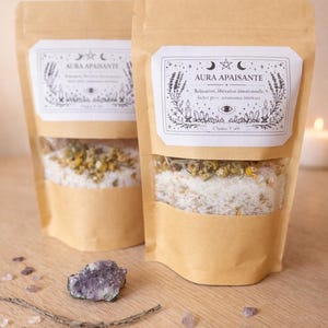 May include: Two brown paper pouches with clear windows, filled with bath salts and dried flowers. Each pouch has a white label with the text "AURA APAISANTE" and decorative illustrations. A purple amethyst and small crystals are on the wooden surface.
