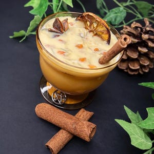 Peut inclure: Un bol en verre rempli d'un dessert crémeux jaune, garni d'une tranche d'orange séchée, d'anis étoilé et d'un bâton de cannelle. Deux bâtons de cannelle sont croisés devant le bol. Des feuilles vertes et une pomme de pin sont en arrière-plan.