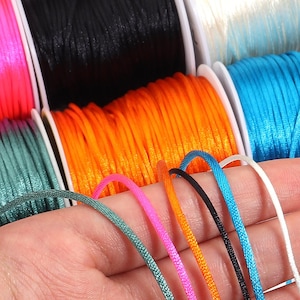 May include: Multiple spools of colourful nylon cord in shades of bright orange, black, white, teal, and neon pink. Several individual strands of the same cord are held in a row, displaying the smooth, braided texture and vibrant colours of the material.