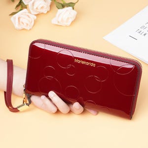 May include: A glossy, burgundy wallet with a zip closure and a wrist strap. The wallet features a pattern of embossed circles and the brand name "Marlekarida" in gold lettering. The wallet is held by a hand.