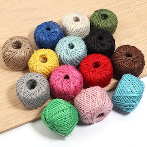 May include: A collection of fifteen small, round balls of twine in various colors including blue, yellow, gray, green, pink, red, brown, black, white, and light blue, each featuring a textured, fibrous surface and a central opening.