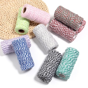 Puede incluir: Una colección de nueve carretes cilíndricos de cordel de panadero, cada uno con un diseño trenzado de dos tonos. Los colores incluyen rosa, verde lima, morado, azul, gris, negro y rojo, todos combinados con blanco en un patrón de rayas.