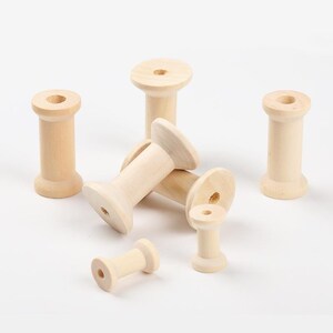 May include: A collection of six unfinished light-coloured wooden spools of varying sizes. Each spool features a cylindrical body with a flared top and bottom, and a central hole running vertically through the middle of the wooden structure.