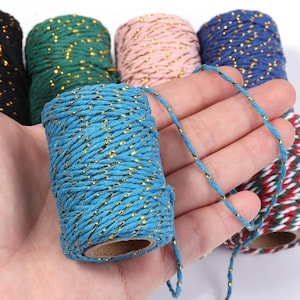 May include: A collection of multi-colored cotton twine spools, each featuring a twisted cord design interwoven with a metallic gold thread. The colors include black, dark green, light pink, royal blue, turquoise, and a red, white, and green stripe.