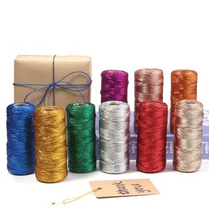 Könnte beinhalten: Eine Sammlung von zehn zylindrischen Garnrollen aus metallischem Kordelmaterial in verschiedenen Farben wie Blau, Gold, Grün, Silber, Rot, Lila, Kupfer und Bronze, dazu ein in braunes Papier gewickeltes Geschenk und ein beiges Etikett mit dem Text thank you.
