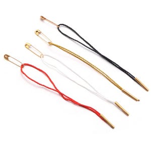 May include: Four garment tags featuring gold-toned safety pins attached to loops of string in red, white, gold, and black. Each string loop is finished with a small, cylindrical gold-toned metal tip at the end, designed for clothing labels.