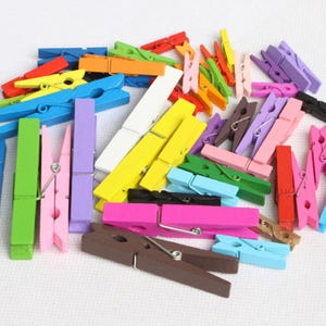 May include: A collection of small wooden clothes pegs in a variety of vibrant colours including red, orange, yellow, green, blue, purple, pink, white, black, and brown. Each clothes peg features a metal spring mechanism for clamping functionality.