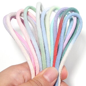 Può includere: Una collezione di molteplici cordini intrecciati in una varietà di colori pastello e vivaci, tra cui rosa, verde chiaro, bianco, azzurro, lavanda e rosso. I cordini sono disposti a ventaglio, mettendo in risalto la loro trama intrecciata.