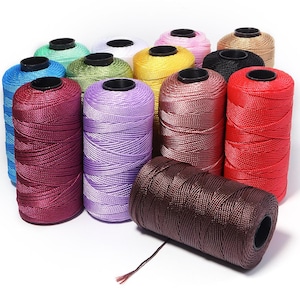 May include: A collection of cylindrical spools of nylon cord in various colors including red, brown, purple, maroon, blue, green, yellow, white, pink, black, and tan. Each spool features a black plastic core and a smooth, tightly wound texture.