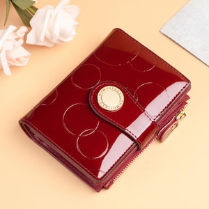 May include: A glossy, claret-coloured wallet with a circular pattern embossed on the front. It has a gold-toned clasp and a zipped compartment. The design is compact and stylish, suitable for carrying essentials.