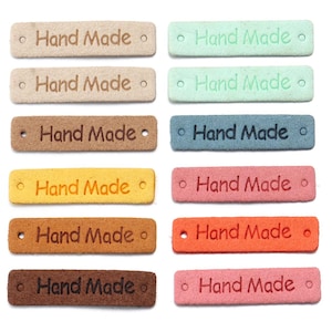 Peut inclure: Une collection de douze étiquettes rectangulaires de différentes couleurs, notamment beige, vert menthe, marron, jaune, rose, orange et brun foncé. Chaque étiquette porte le texte Hand Made en relief au centre et deux petits trous aux extrémités.