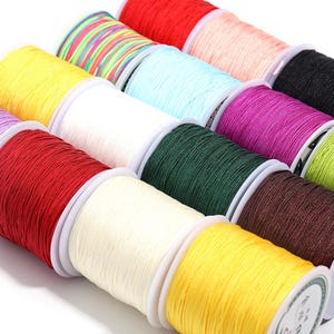 May include: A collection of multiple spools of nylon cord in various solid colors including red, yellow, white, black, green, purple, and brown, alongside a spool featuring a multi-colored striped pattern, all arranged in a grid-like formation.