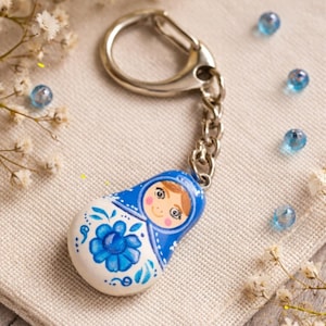 Peut inclure: Un porte-clés avec une breloque lisse en forme de goutte peinte pour ressembler à une poupée russe. La breloque est principalement blanche et bleue avec un motif floral peint à la main et un personnage central aux cheveux bruns.
