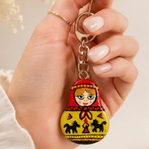 Peut inclure: Un porte-clés avec une petite breloque en forme de poupée russe en bois peinte à la main. La poupée a un foulard rouge, un corps jaune et est ornée de silhouettes de chevaux noirs et de motifs floraux. Elle est fixée à un anneau en métal argenté.