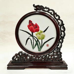 May include: An embroidered artwork in a circular frame, depicting red and green flowers. The dark wood frame is supported by a carved wooden stand. The artwork is a decorative piece, ideal for display.