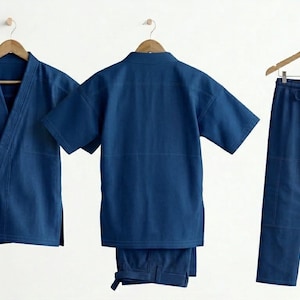 May include: Three-piece indigo blue outfit, including a short-sleeved wrap top with a tie, a short-sleeved top, and pants with a drawstring waist. All pieces are hanging on wooden hangers against a white background.