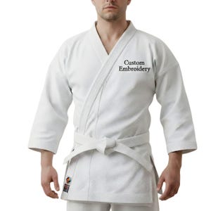 Pode incluir: Uniforme branco de artes marciais com um top envolvente e um cinto a condizer amarrado na cintura. O texto "Custom Embroidery" é visível no peito. O uniforme é feito de tecido texturizado.