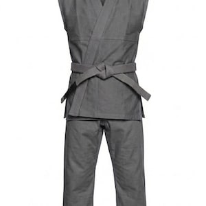 Pode incluir: Um uniforme de artes marciais cinza-escuro sólido composto por uma jaqueta sem mangas com frente transpassada e uma calça comprida a condizer. A jaqueta é presa na cintura com um cinto de tecido atado da mesma cor cinza-escuro.
