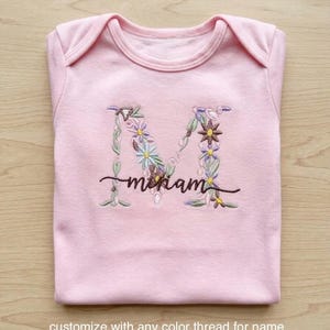 May include: A light pink baby onesie with an embroidered floral design forming the letter "M". The name "Miriam" is embroidered in a cursive font below the letter. The text at the bottom says "customize with any color thread for name".