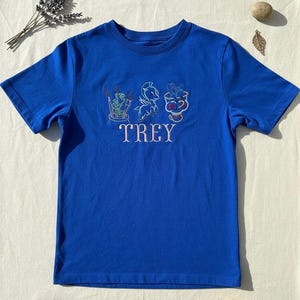 May include: Royal blue t-shirt with embroidered illustrations and the name "TREY" in pink. The embroidery features whimsical designs, including a creature with paintbrushes, a winged horse, and a bowl of fruit. The shirt is made of a soft material.