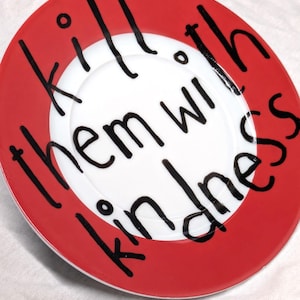 Könnte beinhalten: Ein dekorativer Teller mit rotem Rand und weißer Mitte, auf dem der Schriftzug "kill them with kindness" in schwarzer Schrift steht. Der Text ist kreisförmig angeordnet.