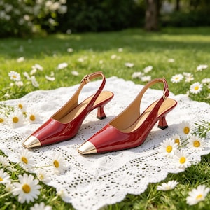Puede incluir: Un par de zapatos de tacón slingback de charol rojo con punteras cuadradas doradas. Los zapatos tienen un tacón pequeño y correas de tobillo ajustables. Los zapatos se exhiben sobre un paño de encaje blanco, rodeados de margaritas.