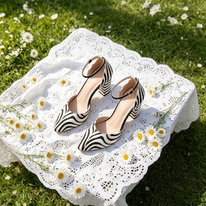 Könnte beinhalten: Ein Paar zebra-gestreifte High Heels mit Knöchelriemen. Die Schuhe haben einen Blockabsatz und eine spitze Zehenpartie. Sie liegen auf einer weißen Spitzentischdecke, mit Gänseblümchen verstreut.