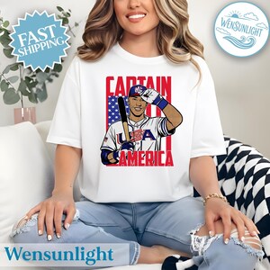 May include: White t-shirt featuring a baseball player with a bat, wearing a USA uniform. The design includes the text "CAPTAIN AMERICA" in red, with a background of the American flag. The shirt has a round neck.