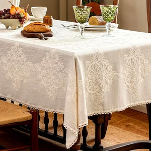 Puede incluir: Un mantel crema con intrincados detalles de encaje cubre una mesa de madera. El mantel presenta un borde festoneado y está adornado con un patrón floral. La mesa está puesta con cristalería, pan y un cuenco de fruta, creando una elegante escena de comedor.