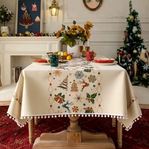 Peut inclure: Une nappe rectangulaire de couleur crème ornée d'un motif central vertical composé de sapins de Noël, de rennes, de flocons, de cloches et de cadeaux colorés, finie par une bordure à pompons blancs, disposée sur une table en bois.