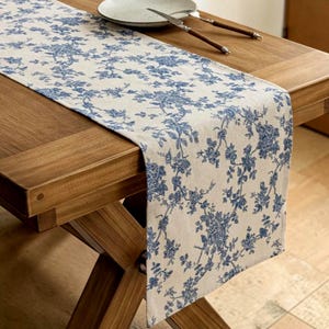 Puede incluir: Camino de mesa rectangular con fondo crema y estampado floral azul. El camino de mesa está colocado sobre una mesa de madera, con un plato y cubiertos visibles. El diseño floral presenta flores y hojas azules detalladas.