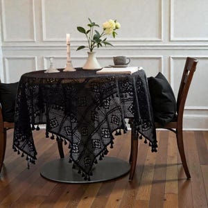 May include: A black crocheted tablecloth with a geometric pattern and small tassels along the edges, draped over a round table. The table holds a white vase with yellow flowers, a candle in a white holder, and a grey mug on a book.