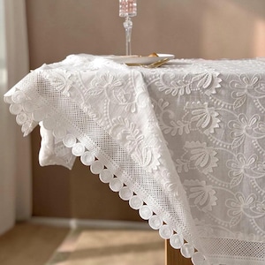 May include: A white lace tablecloth with detailed floral embroidery and a scalloped edge. The tablecloth is draped over a wooden table, highlighting the delicate details and texture of the fabric. A glass and plate are on the table.