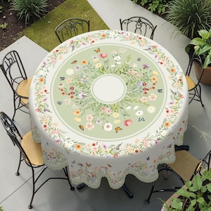 Puede incluir: Un mantel redondo con un diseño floral y de mariposas. El mantel presenta un centro verde claro con un círculo de flores y mariposas. El borde tiene un borde festoneado y un patrón floral repetitivo. Sillas de hierro forjado rodean la mesa.