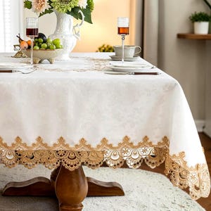 Pode incluir: Uma toalha de mesa branca com borda de renda dourada, sobre uma mesa de madeira. A toalha de mesa tem um padrão floral sutil. Taças de vinho, um vaso de flores e frutas adornam a mesa, sugerindo um ambiente de jantar.