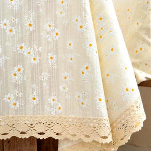 Peut inclure: Nappe crème avec un motif de marguerites. Les marguerites blanches ont des centres jaunes. Le bord de la nappe est orné d'une bordure en dentelle beige. Le tissu a un aspect texturé.
