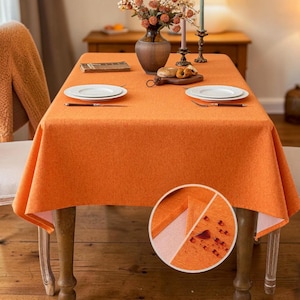 May include: A rectangular orange tablecloth with a textured fabric finish draped over a dining table. The surface features two white plates, a dark vase with dried flowers, and a wooden board with bread. An inset shows water droplets.