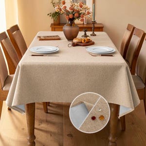 May include: A rectangular beige tablecloth with a textured fabric finish draped over a wooden dining table. The tablecloth features a water-resistant surface, shown in a circular inset with three liquid droplets resting on the fabric material.