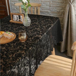 Puede incluir: Un mantel de encaje negro con un intrincado patrón floral de damasco sobre una mesa. En la superficie hay un jarrón de cristal transparente con flores blancas, un calendario perpetuo de madera que muestra OCT 3 y un plato de madera con un pastel.