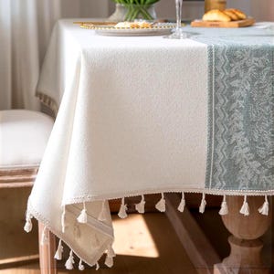 Pode incluir: Uma toalha de mesa retangular com um centro de cor creme sólida e uma borda verde sálvia com um padrão botânico detalhado. As bordas são finalizadas com pequenas borlas de tecido cor creme penduradas ao longo de todo o perímetro.