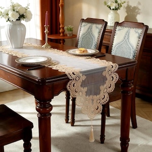 Pode incluir: Um caminho de mesa retangular feito de tecido branco transparente com bordas de renda bege intrincadas com padrões florais e bordas recortadas. O caminho pende das extremidades de uma mesa de madeira escura e inclui uma borla decorativa.